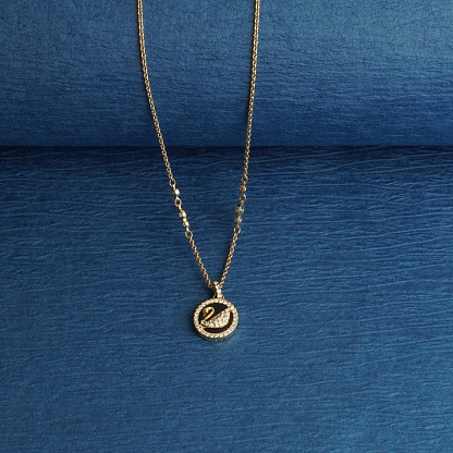 The Necklace has a gold chain and a pendant. The pendant is circular shaped with design of black and gold Swan. It has small white diamonds studded on the periphery. Close up on dark blue background.