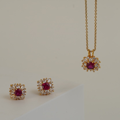 A set of one necklace and two earrings with square shaped big red ruby in the centre and white shining small diamonds around it on the periphery. Necklace has a gold chain and pendant. Close up on White background. Necklace is hanging and earrings are placed on white table.