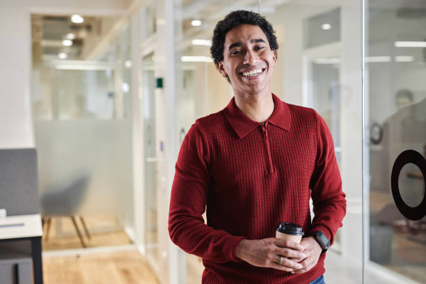 istock Young Arabian man holding coffee cup standing by glass wall in office 1631428088