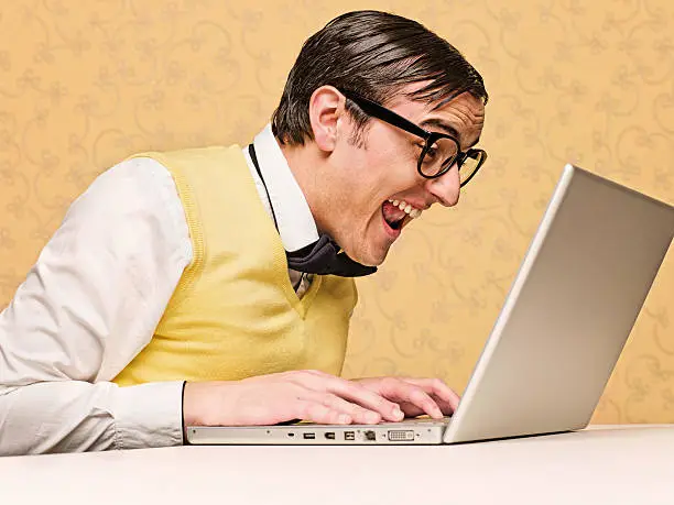 Young male nerd sitting at the computer, he is laughing out lound, and watching the screen.