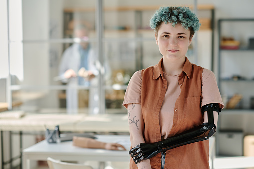 Waist up portrait of young woman with bionic prosthetic arm posing in doctors office and smiling at camera, copy space