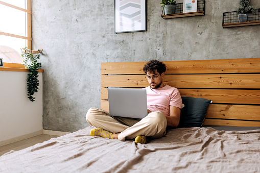Young casually clothed man using laptop in bedroom at home