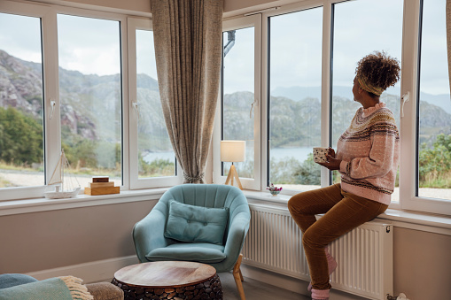 A medium wide angle view of a mature woman on her own sitting in the living room of a staycation holiday rental. She is sitting on the windowsill and looking out along the coast of Loch Torridon in the Scottish Highlands. She is holding a warm mug with a hot drink.