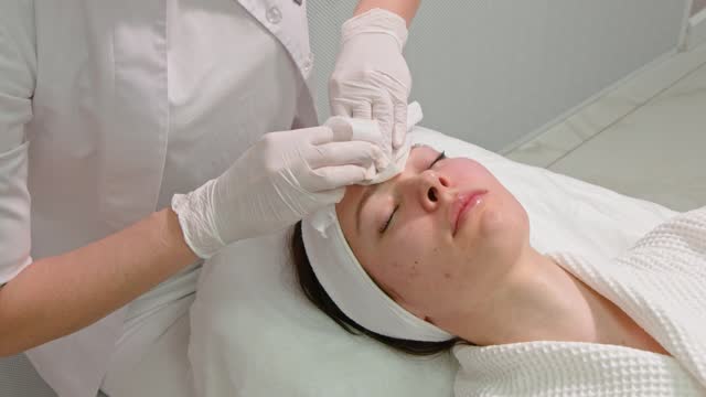 A beautician applying a mask to the face of a young reclining woman in a beauty salon for a skin cleansing and moisturizing procedure.