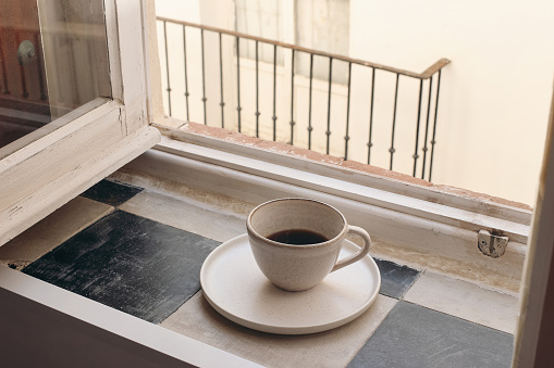 Cup of coffee on vintage in ceramic mug checkered tile windowsill. Blurred beige wall with balcony background. Calm lifestyle scene with hot drink. Breakfast, relaxation concept, no people.