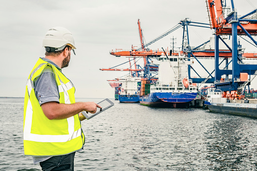 Dockworker standing in front of a container terminal in port while checking his digital tablet computer. The man is wearing protective workwear and looking at the tablet.