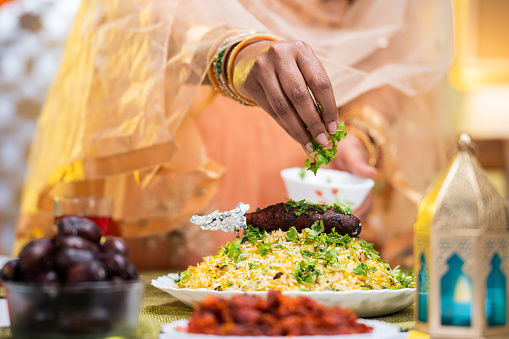Close up shot of indian woman hands adding toppings or coriander on top of biryani at home - concept of iftar dinner preparation, festival celebration and Ramadan feast