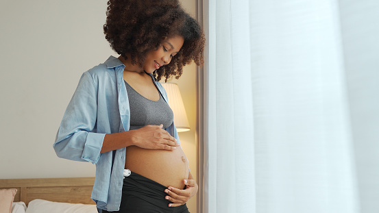Portrait of Black African American Pregnant woman in curly hair standing embrace her belly at home. Expecting mother. Pregnancy concept.