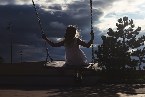 Silhouette of little girl swinging on swing in urban park at cloudy sky background. Rear back view of kid 5-6 year old swinging in sunshine playground outdoors. Bad weather concept. Copy ad text space