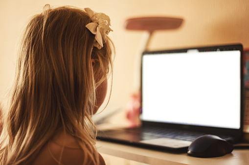Back view of little girl child 5-6 year old looking laptop in child room watching on empty isolated screen. Adorable girl kid education online at home. Childhood education concept. Copy ad text space