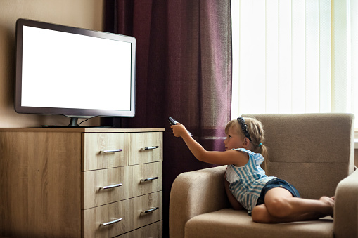 Little girl child 5-6 year old lying down on chair with remote control watching tv, empty isolated screen. Cute girl kid looking cartoon on tv channel. Television addiction concept. Copy ad text space