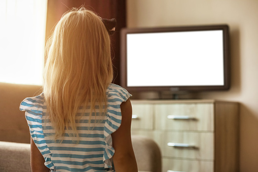 Rear view of little girl kid 5-6 year old watching looking tv in living room looking movie. Adorable addiction girl child, empty isolated screen. Television dependency tv concept. Copy ad text space
