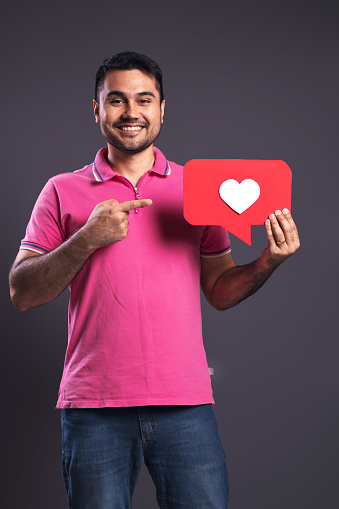 Portrait of a Brazilian wearing a pink polo shirt, holding with his left hand a speech bubble with a white heart inside, with his right hand pointing to it, happy, looking at the camera and smiling - Belém - Pará - Brazil