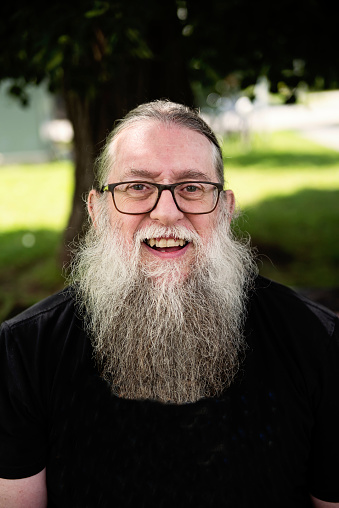 Portrait of a senior man with a long beard outdoors in summer. He is wearing glasses and looking at the camera with a big smile. Vertical head and shoulder outdoors shot with copy space. This was taken in Montreal, Quebec, Canada.