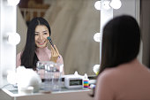 Cute young Asian woman looking at herself in the mirror while applying makeup.