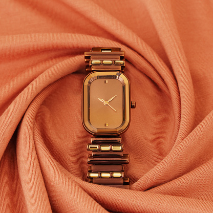 A beautiful ladies wrist watch. The watch face or dial is rectangular. It is dark brown in colour. Watch belt is made up of cylindrical links of dark brown and gold colours. The watch is placed on a spiral arrangement of a dark peach fabric. Watch face has hour and minute hands that show 10 minutes past 10 o'clock or 10:10.