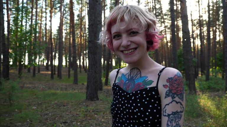 Carefree hippie woman with tattoos going along path at forest. Happy smiling punk girl with pink hair looking into camera outdoor. Female hipster enjoying stroll outdoor admiring beautiful scenic