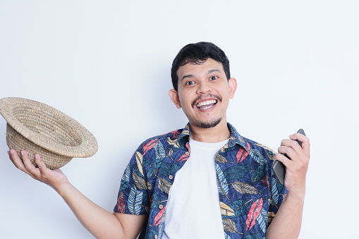 Asian man wearing beach shirt smiling happy while holding mobile phone and straw hat