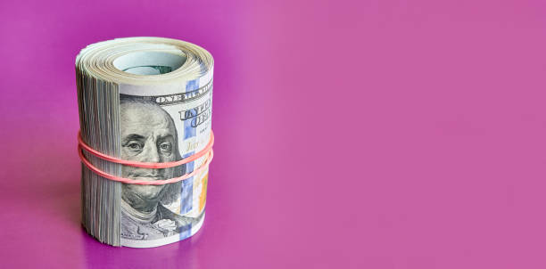 istock Many hundred dollar bills rolled up with a red rubber band on a pink background. 1631828706