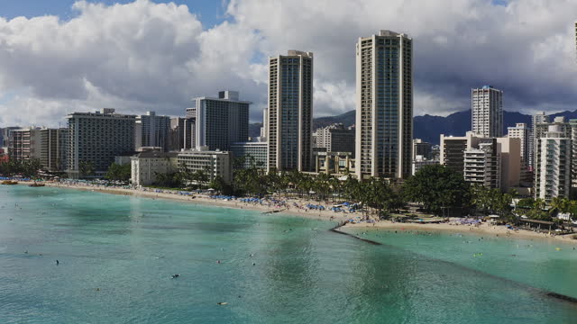 Aerial view of Waikiki beach high-rise hotels and resorts with dramatic clouds, surfers catching smooth ocean waves in warm afternoon light