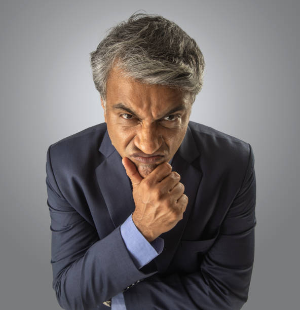 High angle portrait of angry senior businessman touching chin and staring at camera while standing against white background