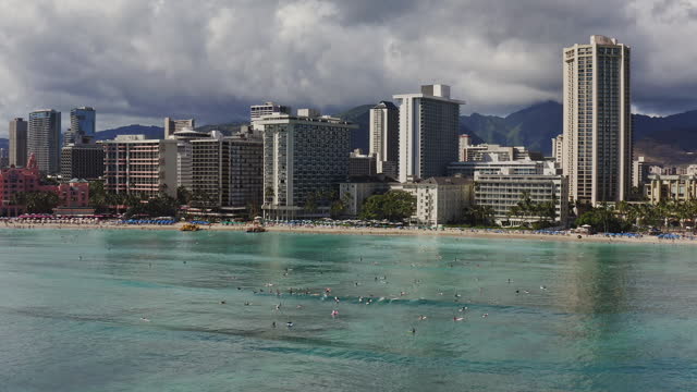 Aerial view of Waikiki beach high-rise hotels and resorts with dramatic clouds, surfers catching smooth ocean waves in warm afternoon light