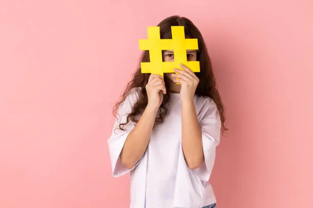 Photo of Little girl covering face with social media hashtag symbol, recommending to follow trendy content.