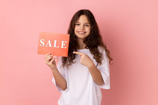 Portrait of joyful adorable little girl wearing white T-shirt pointing at card with sale inscription, looking at camera with toothy smile. Indoor studio shot isolated on pink background.