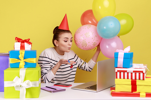 Grateful woman sitting workplace surrounded by gift boxes and balloons, sending air kisses to laptop screen, celebrating birthday by video call. Indoor studio studio shot isolated on yellow background