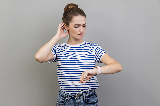 Portrait of pensive thoughtful woman wearing striped T-shirt standing looking at her wrist watch, having confused puzzled expression. Indoor studio shot isolated on gray background.