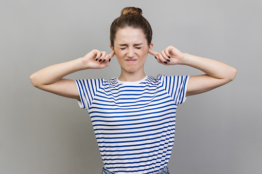 I dont want to hear this. Annoyed frustrated woman wearing striped T-shirt closing ears with fingers ignoring information, tired of irritating noise. Indoor studio shot isolated on gray background.