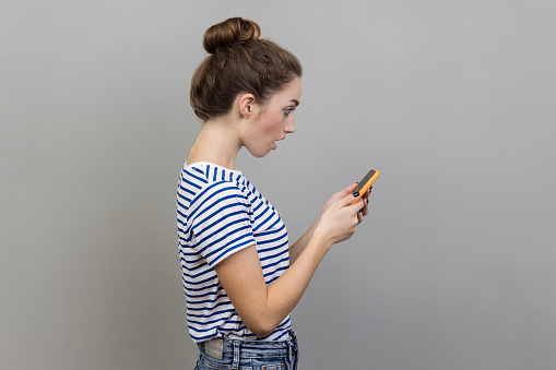Side view of beautiful astonished woman blogger wearing striped T-shirt using smart phone, looking at device screen, sees shocking content. Indoor studio shot isolated on gray background.