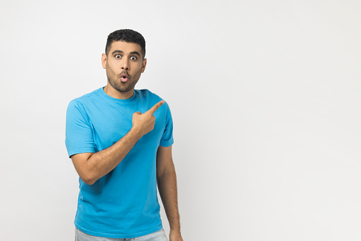 Portrait of shocked astonished unshaven man wearing blue T- shirt standing pointing copy space for advertisement or promotional text. Indoor studio shot isolated on gray background.