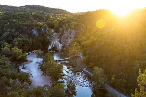 Aerial view of water cascading over rocks into a natural pool at Turner Falls in Oklahoma on sunset, beautiful nature, water, road and rocks among trees.
