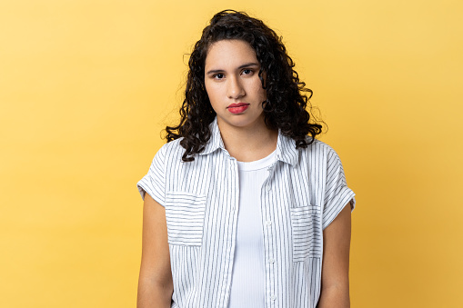 Portrait of sad desperate woman with dark wavy hair standing looking at camera, being in bad mood, suffering depression. Indoor studio shot isolated on yellow background.