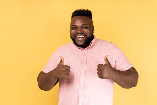 Portrait of pleased optimistic smiling man wearing pink shirt showing thumbs up gesture, doing success sign with hand, approved excellent job. Indoor studio shot isolated on yellow background.