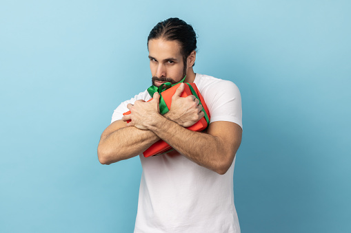 Long-awaited present. Portrait of greedy handsome man with beard wearing white T-shirt embracing red gift box, looking at camera. Indoor studio shot isolated on blue background.