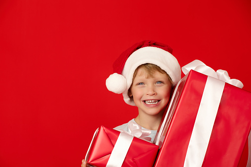 Large boxes with gifts were presented by Santa Claus to little boy in New Year's hat with pompom. In big red hat, child stands among gift boxes, smiling, on red background