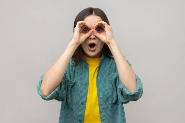 istock Woman standing with hands on eyes binoculars gesture and looking at camera with surprised face. 1631878089