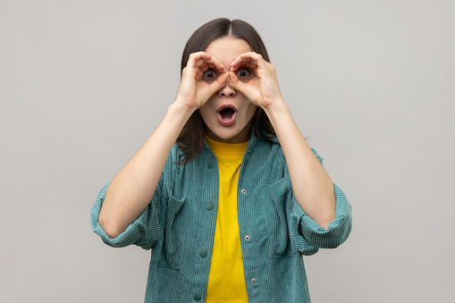 Shocked attractive young woman standing with hands on eyes binoculars gesture and looking at camera with surprised face, wearing casual style jacket. Indoor studio shot isolated on gray background.