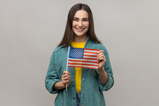 Portrait of young happy satisfied woman standing and holding american flag, celebrating national holiday, wearing casual style jacket. Indoor studio shot isolated on gray background.