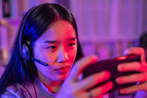 Portrait of Young Asian Woman smile while use smartphone to playing online game in neon lights room and use headphone to listening and talking during competition with excited