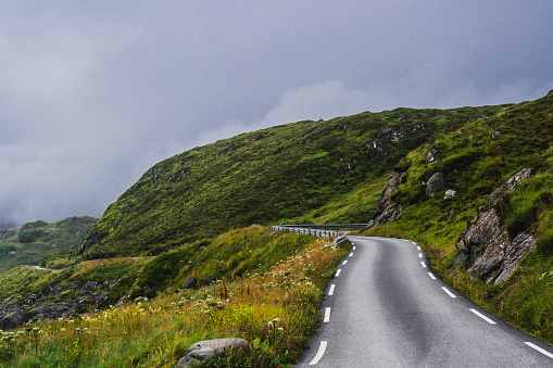 A horizontal landscape photograph shows a wet road in Norway near Maløy, on the mountainside. The scene features mountains blanketed with lush green vegetation and areas with stones, under a cloudy sky. Captured during a summer morning, the image evokes a sense of tranquility and connection to nature.