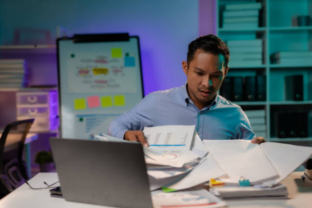 istock Serious young Asian male worker working alone with computer and documents in office at night. Focused worker busy managing deadlines staying late at work. Workaholic concept. 1631894213