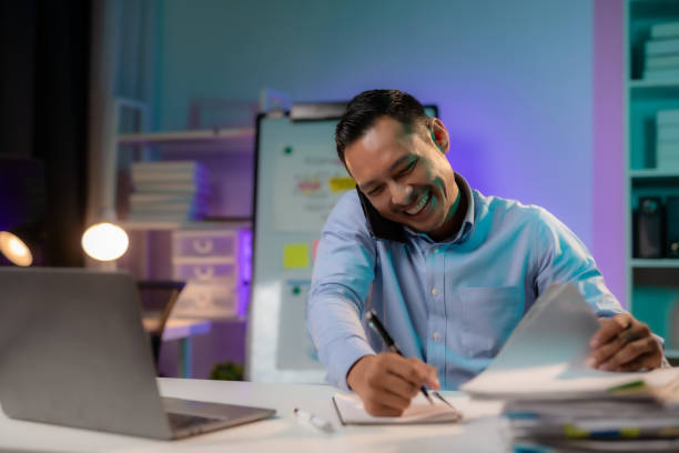 istock Late-Night Hustle. Young Asian Business Professional Talking on Phone, Putting in Extra Work. Ambitious Asian Professional. Overtime Work, Focused Phone Talk in Corporate. 1631894635