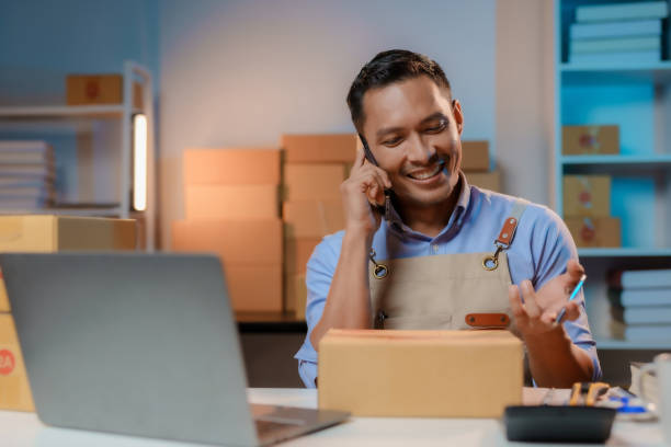 istock Overtime work concept. Asian male shop owner preparing parcels for boxes writing customer addresses and talking on the phone. 1631895169
