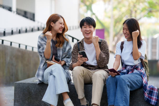 group of asian students rejoice with their arms raised in joy after receiving the results of their university entrance exams via their mobile phones. - news of the world imagens e fotografias de stock