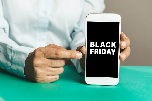 Unrecognizable person is showing screen of smart phone to camera over green table and is pointing with one finger at device screen in front of gray background. Black Friday text on screen.
