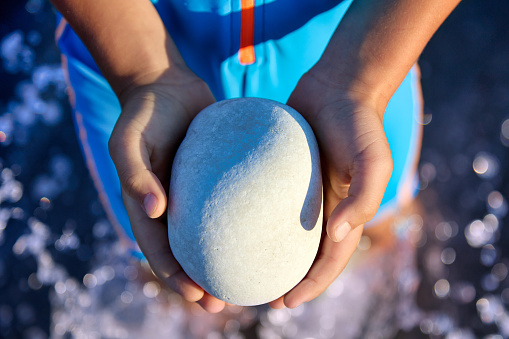 Top view of a smooth gray stone in the hands of a small child on the beach. Close-up of a heavy stone that a child holds in his palms while standing in the water shows to the camera