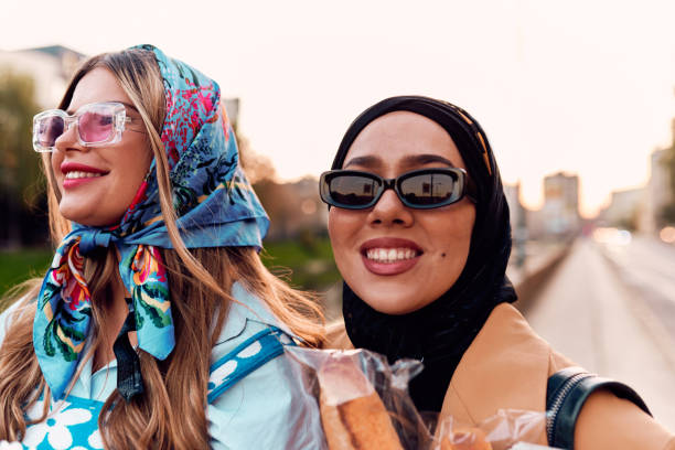 Couple woman one wearing a hijab and a modern yet traditional dress, and the other in a blue dress and scarf, walking together through the city at sunset. One carries a bouquet and bread, while the other holds a blue handbag, symbolizing a beautiful fusion of cultures, friendship, and serene urban charm. pareja de mujeres una con un hijab y un vestido moderno pero tradicional, y la otra con un vestido azul y bufanda, caminando juntas por la ciudad al atardecer. uno lleva un ramo y pan, mientras que el otro sostiene un bolso azul, que simboliza un hermoso f